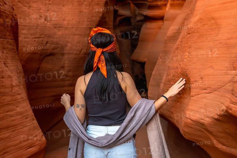Woman with an orange bandana and gray scarf hiking through the narrow slot canyon, touching the vibrant orange red sandstone walls, finding adventure in nature