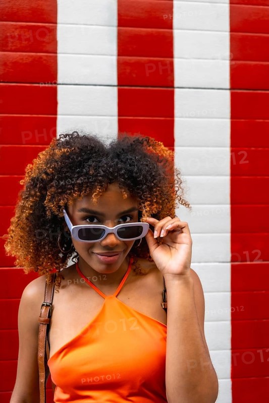 Confident young woman posing with style, adjusting her sunglasses with a playful expression against a vibrant red and white striped background, showcasing fashion and positive lifestyle