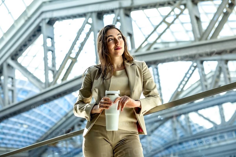 Young businesswoman with a hopeful expression holding her reusable travel mug while contemplating future business opportunities in a modern corporate setting with sleek architecture