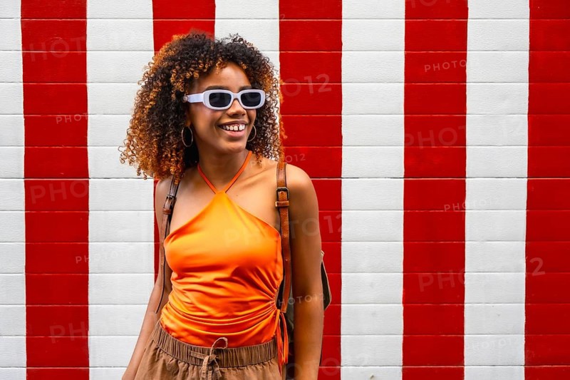 Joyful young woman in sunglasses and an orange top poses against a bold red and white striped wall, exuding confident, colorful urban summer style and carefree energy