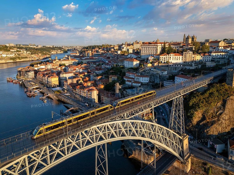 Drone view of Dom Luis I Bridge over Douro River in Porto, Portugal, with riverside quay, including a tram on the bridge and rabelo boats by the quay at sunrise