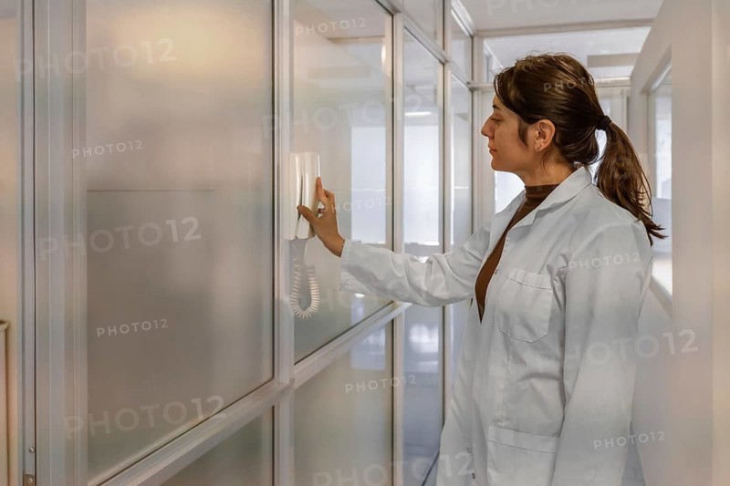 Young female scientist in white lab coat using a wall-mounted intercom handset in a bright institutional corridor with frosted glass partitions and clean white walls