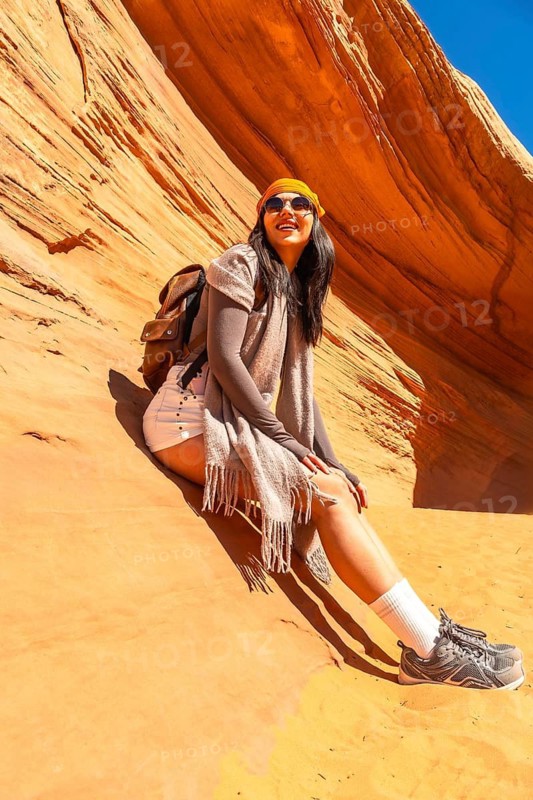 Young woman with a backpack and sunglasses smiling while sitting on the sandstone ground inside biidi the arch cave, enjoying a sunny day exploring the unique slot canyon landscape