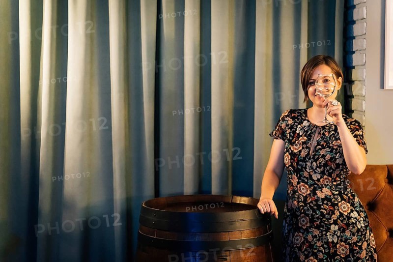 A woman closes her eye with a wine glass standing near a wine barrel. A woman in a dark floral dress stands in a bar interior holding a wine glass near her face beside a wooden barrel. Warm lighting, tall curtains, and rustic decor create a cozy hospitality atmosphere suitable for wine tasting, bar service, or restaurant promotion
