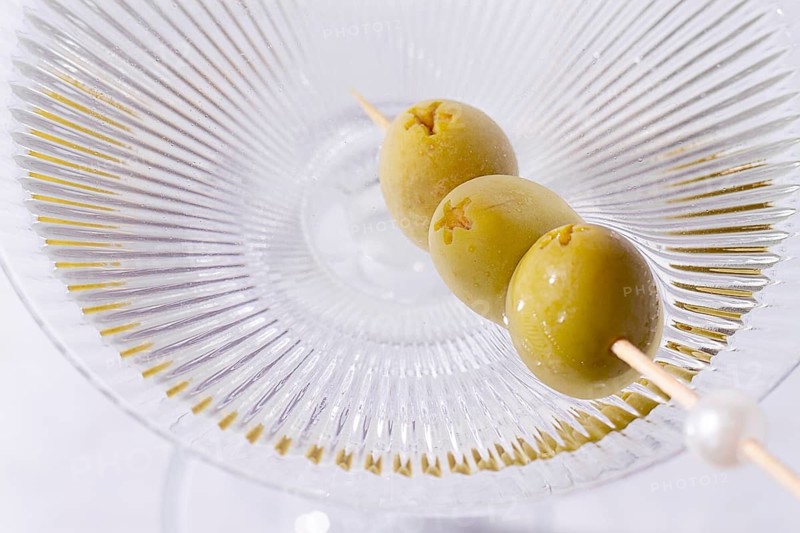 A clear glass cocktail sits on a white marble surface. Three green olives are placed on a skewer resting inside the glass. The light shines on the glass, highlighting its shape