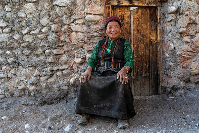 Friendly elderly woman in traditional clothing sitting in front of wooden door, Tangge, Mustang, Upper Mustang, former kingdom, Nepal