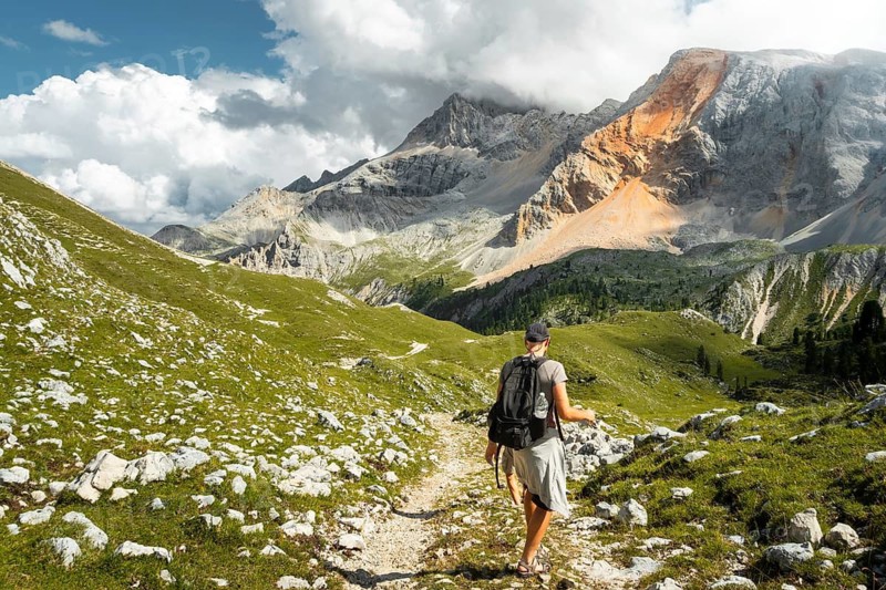 Male hiker with backpack walking dirt trail green alpine meadow majestic mountain peaks background Malga Rossalm Dolomites Italy summer adventure travel lifestyle