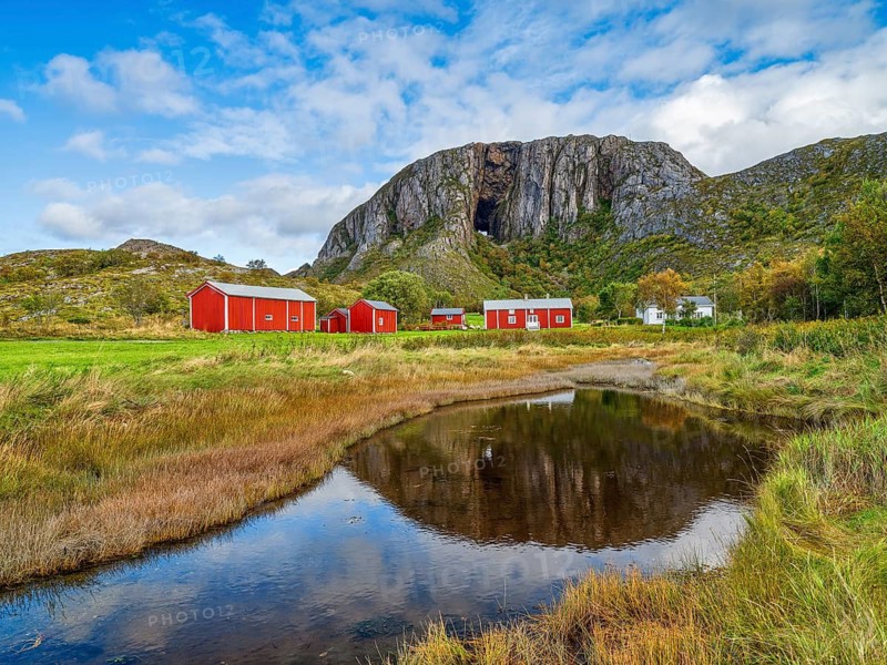 Red buildings against a mountain backdrop with sky and clouds, a quiet pond in the foreground, Torghatten, mountain, Brønnøysund, Helgelandskysten, Norway