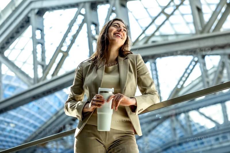 Woman in a professional suit holding a white reusable coffee cup and looking up, smiling in a bright, modern architectural setting, embodying a successful and sustainable urban lifestyle