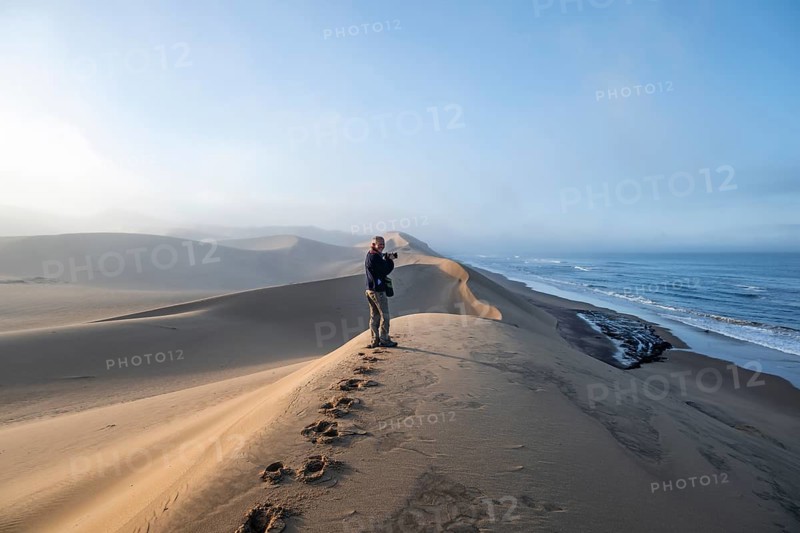 Tourist, photographer on a sand dunes in morning light, view of Atlantic coast and sea, Sandwich Harbour, impressive desert landscape, Namib Naukluft Park, Namibia