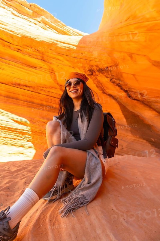 Young woman smiling and sitting on a textured orange sandstone rock formation, exploring the beautiful and unique natural wonder of biidi the arch cave in page, arizona, usa