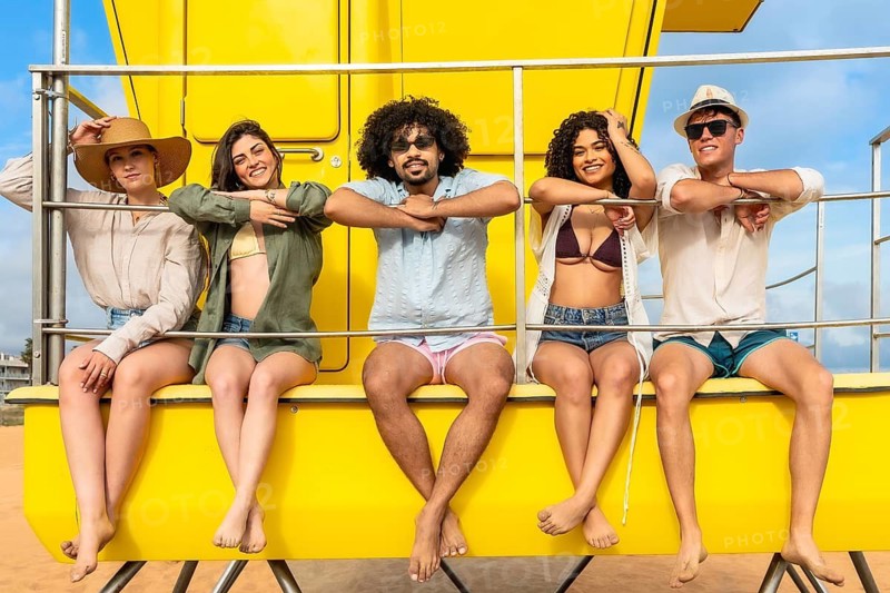Diverse group of young adult friends enjoying summer vacation at the beach, sitting barefoot and smiling on a bright yellow lifeguard tower representing freedom and connection