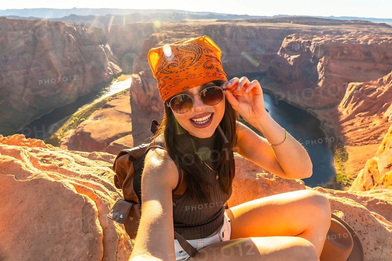 Young woman smiling and adjusting sunglasses, sitting on sandstone cliffs overlooking the colorado river in famous horseshoe bend, experiencing a desert adventure