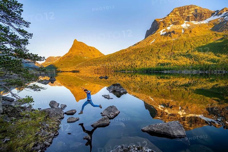 Young man jumping from stone to stone in water, picturesque mountain landscape in the Innerdalen high valley with Innerdalstårnet summit, mountain peaks reflected in Lake Innerdalsvatna, in the evening light at sunset, Trollheimen Mountain Area, Sunndal, Møre og Romsdal, Norway