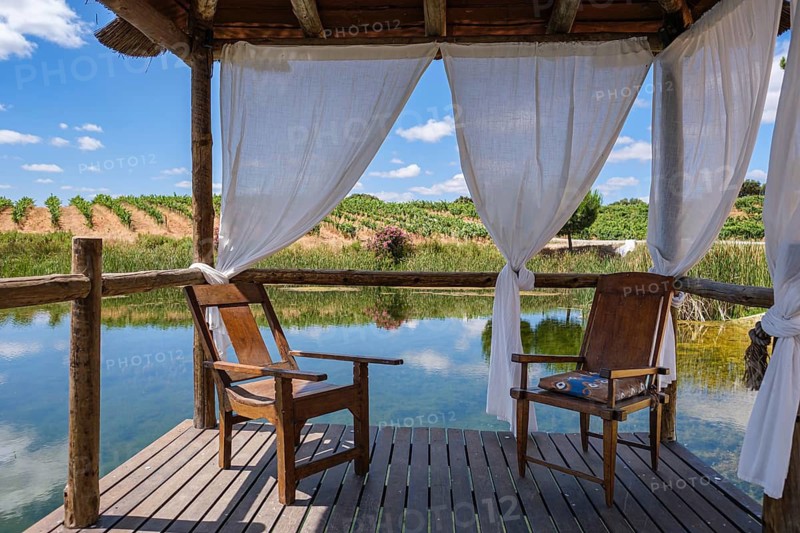 Two wooden chairs in a pavilion with curtains standing over a calm lake, Pedrógão, Beja, Portugal