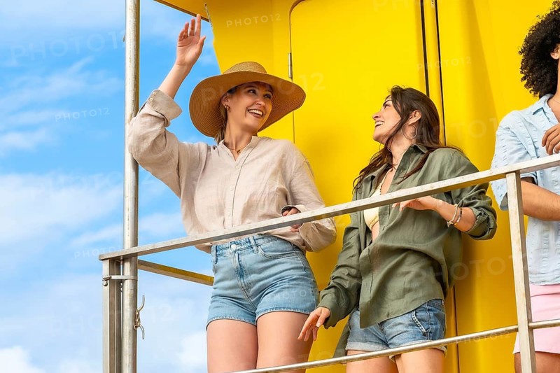 Group of diverse happy young women enjoying sunny summer vacation, standing on a bright yellow balcony with a clear blue sky, laughing and smiling while having fun outdoors