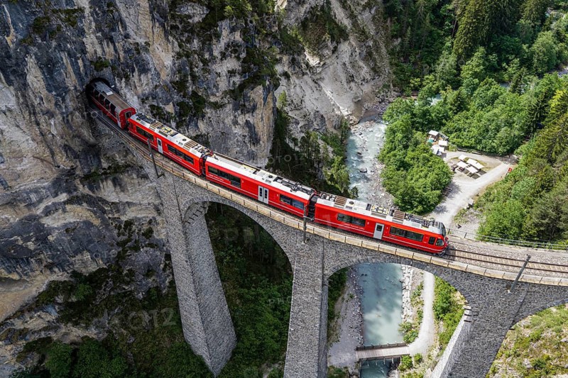 Red train of the Rhaetian railroad line, Bernina Express, passing the Landwasser Viaduct, arch bridge over Landwasser river, aerial view, Switzerland