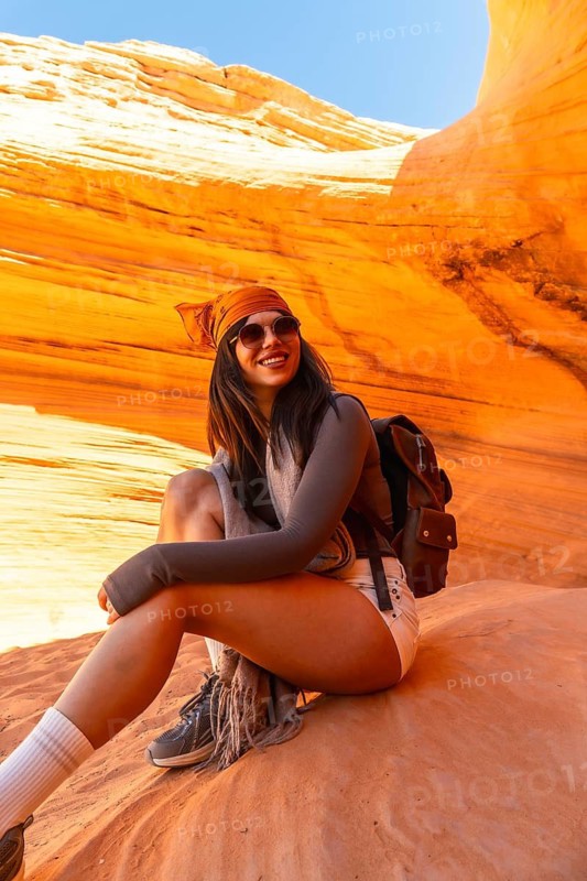 Happy woman wearing backpack and bandana sitting on carved sandstone rock inside biidi the arch cave, enjoying a moment of rest during her desert adventure