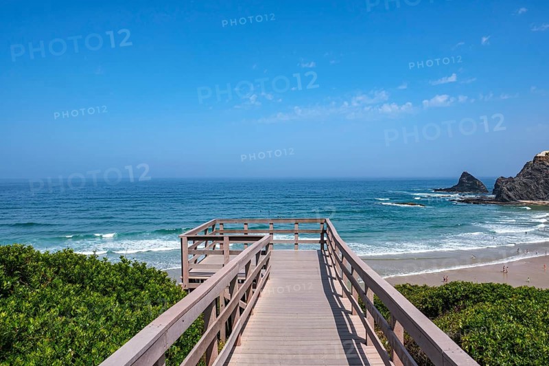 A wooden walkway with views of the sea and rocks under clear skies surrounded by vegetation, Odeceixe, Faro, Portugal