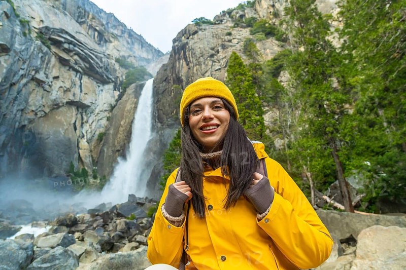 Woman hiker wearing a yellow jacket and beanie smiling at the camera, enjoying the natural beauty of lower yosemite falls and surrounding mountains in yosemite national park