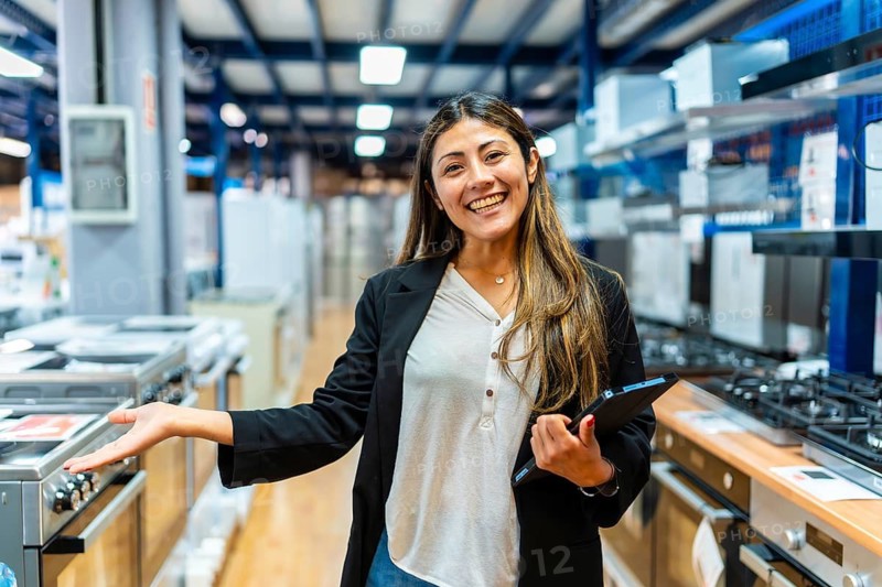Confident saleswoman smiling while holding a digital tablet and gesturing towards kitchen appliances in a modern retail showroom, assisting customers with home renovations