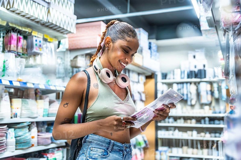 Young woman smiling while browsing selection of products in a retail store aisle, wearing headphones around her neck and considering a purchase
