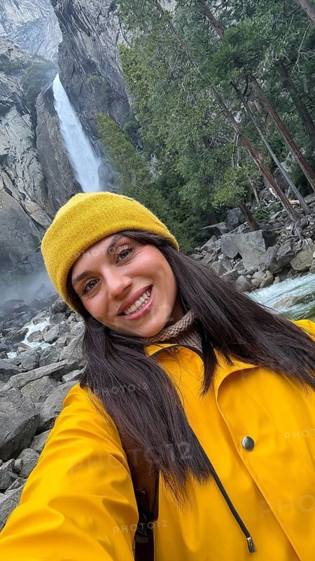 Woman in a yellow jacket and beanie smiles for a selfie while hiking at yosemite national park, enjoying the scenic lower yosemite falls, rocks, trees and rushing water