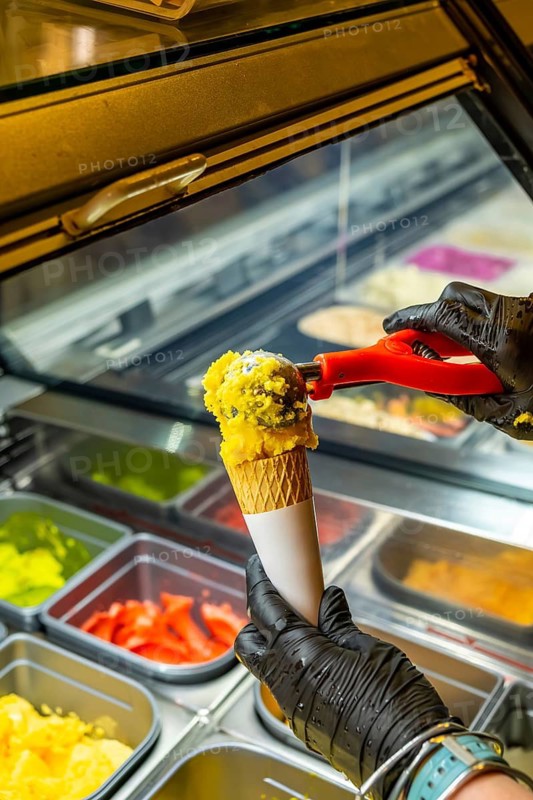 Person wearing black latex gloves serving creamy yellow ice cream with sprinkles into a crispy waffle cone from a display refrigerator filled with various vibrant flavors
