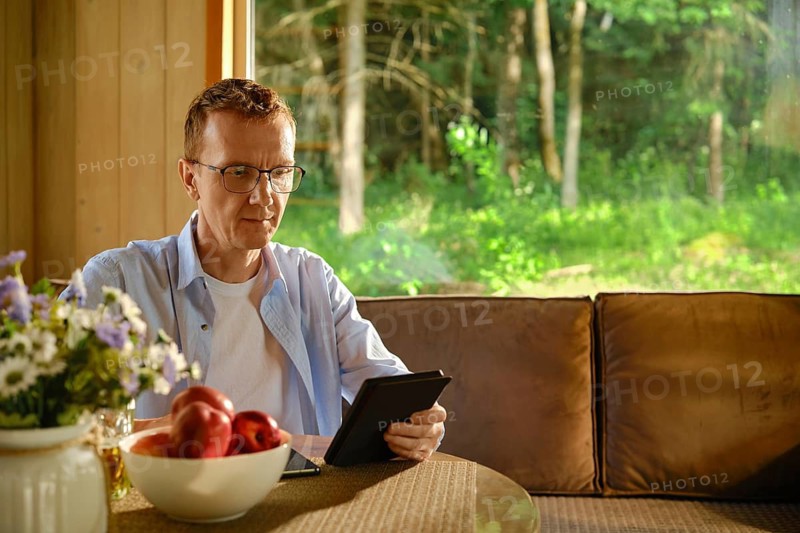 Middle-aged man in glasses sits at table in wooden cabin with e-book. Sunlight enters room through large window creating bright atmosphere