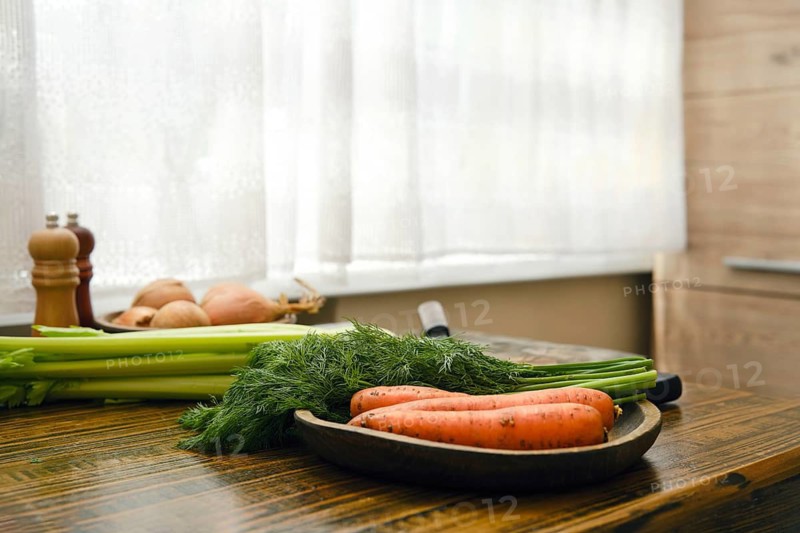 Vegetables are on a wooden table in a kitchen. Carrots, dill and celery are arranged neatly on a plate. The sun shines through the window, creating a bright environment