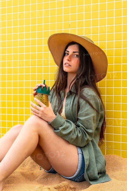 Woman wearing straw hat and linen shirt sitting on sand against yellow tiled wall, holding a pineapple shaped drink bottle and relaxing in a summer setting