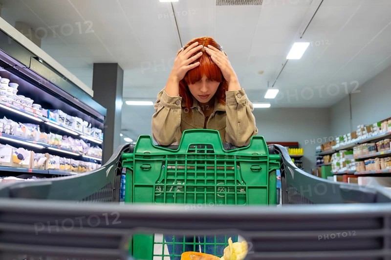 Woman feeling overwhelmed, holding her head in a grocery store, struggling with the financial pressure of high inflation and increasing cost of living reflected in her almost empty shopping cart