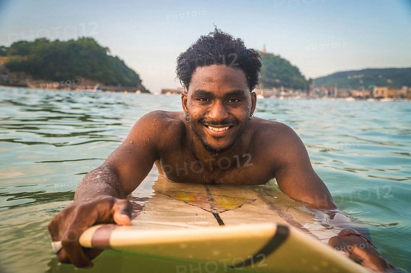 Smiling young black male surfer relaxing on his surfboard in the ocean during a summer day, enjoying lifestyle and water sports
