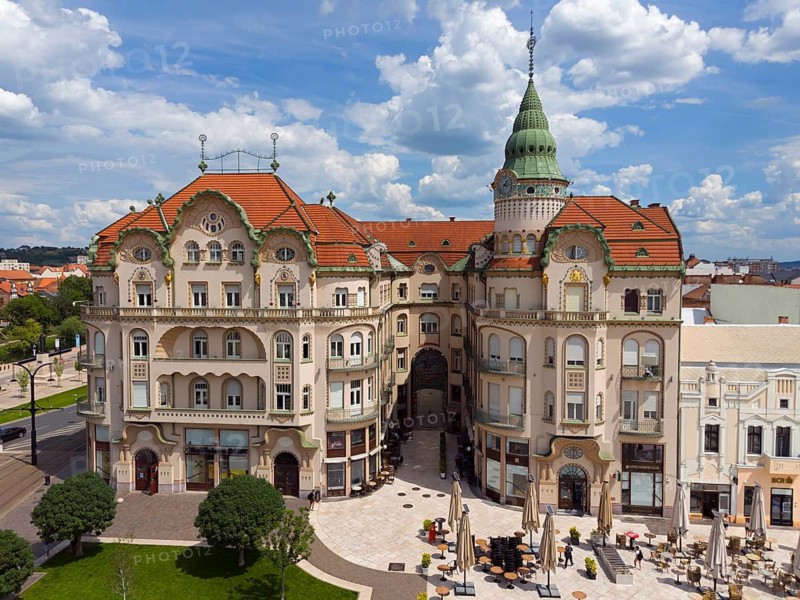 Large baroque building with red roof and green tower in a busy square under cloudy sky, aerial view, Black Eagle Palace, Piata Unirii, Unity Square, Oradea, Oradea, Oradea, Bihor district, screechland, Transylvania, Romania