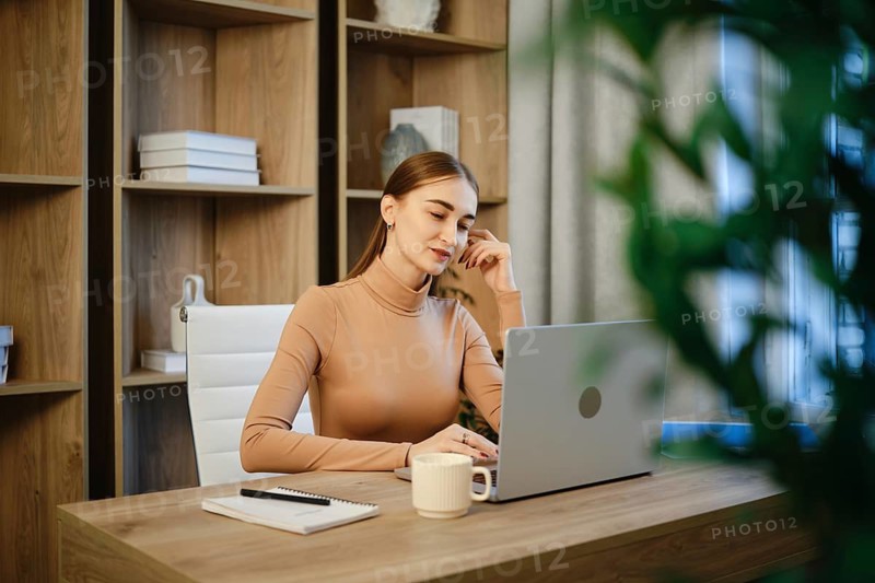 A woman sits at a wooden desk in a home office. She is focused on her laptop. A notebook and a cup are beside her. Plants and shelves are in the background