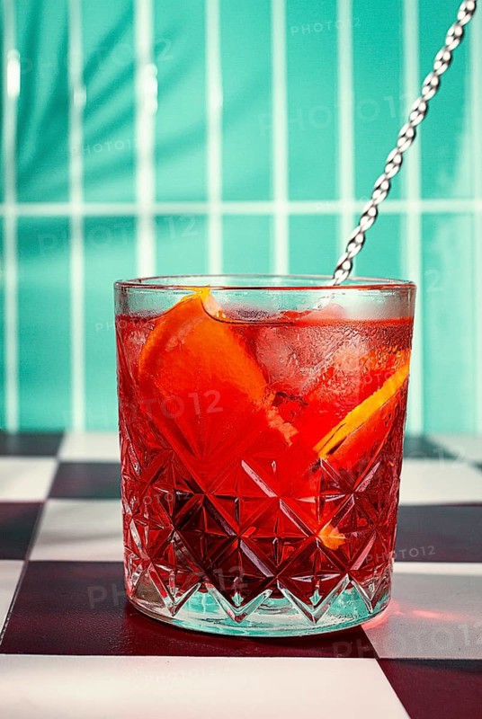 A cocktail with orange slices rests in a glass on a black and white checkered table. The background features green tiles and is bright and colorful