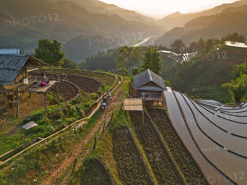 Aerial view of woman with pink backpack sitting on a wooden house at sunset in the mountains and rice terraces just in front of setting the new trip at Mù Cang Ch?i, Mu Cang Chai District, Lào Cai Province, North Vietnam, Vietnam