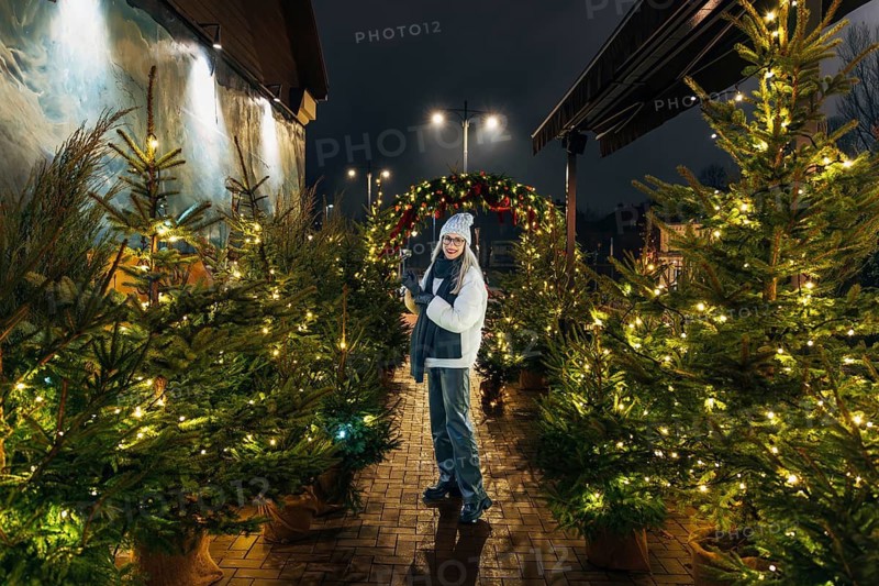 Young woman enjoying festive christmas tree display at night. A woman wearing a white winter hat, scarf and jacket stands outdoors at night holding a camera among Christmas trees decorated with warm yellow lights. Behind her, a festive arch adorned with greenery and red ornaments adds depth to the illuminated holiday setting. Streetlights and decorative lighting create a cozy seasonal atmosphere, combining winter attire, glowing decorations and nighttime city ambience