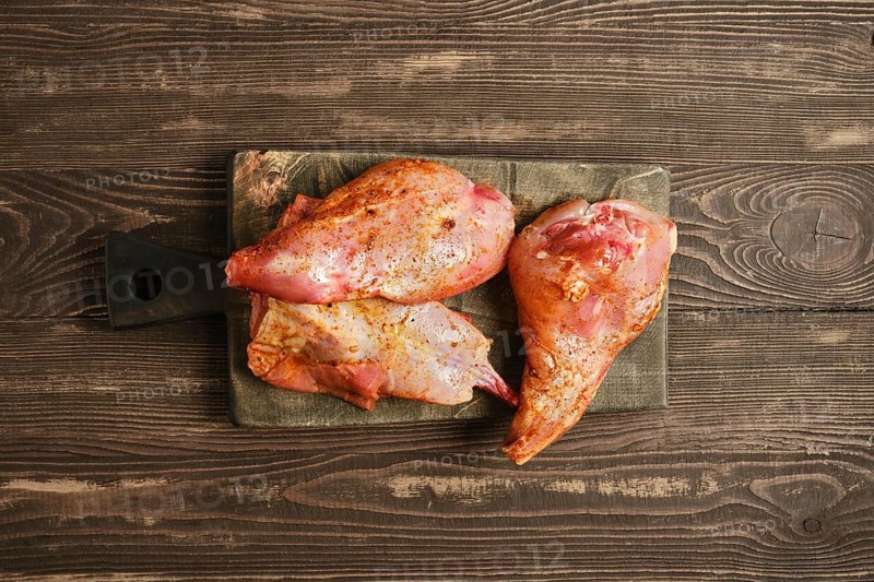 Top view of marinated rabbit meat on a wooden cutting board. They show signs of seasoning. The kitchen countertop is simple. A knife is nearby, suggesting preparation for cooking
