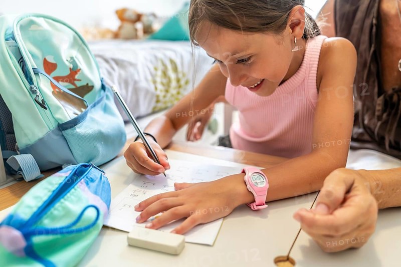 Happy granddaughter smiling while studying and writing numbers on paper, with school supplies on the table, receiving assistance from her grandmother during her education