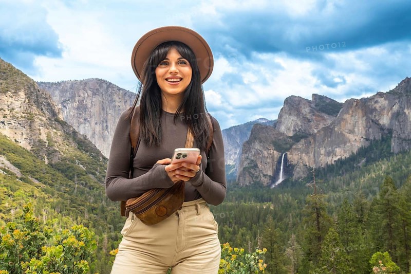 Woman smiling, holding a smartphone, and standing in front of the impressive yosemite national park landscape, enjoying the stunning view of tunnel view, bridalveil falls, and el capitan