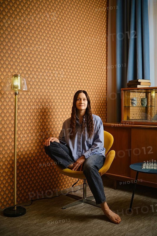 Woman with long hair sits in yellow chair at home. Wall has unique pattern. Floor lamp stands beside her. A display case with items is visible behind her. Chess set is on table in front