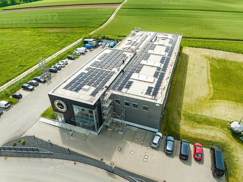 Building with solar panels on the roof, surrounded by fields and parked cars, PV assembly, Perrot company, Calw, Germany