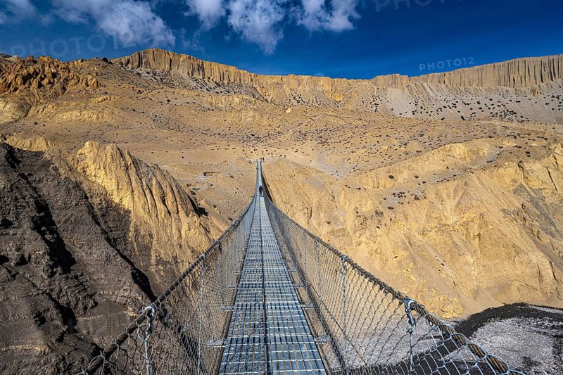 A man walks over a very long suspension bridge made of steel cables across a river valley, mountain landscape, Mustang, Upper Mustang, former kingdom, Himalayas, Nepal
