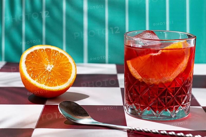 A clear glass holds a colorful drink with slices of orange. Next to it, there is a halved orange on a checkerboard table. The scene is bright and lively in a sunny location