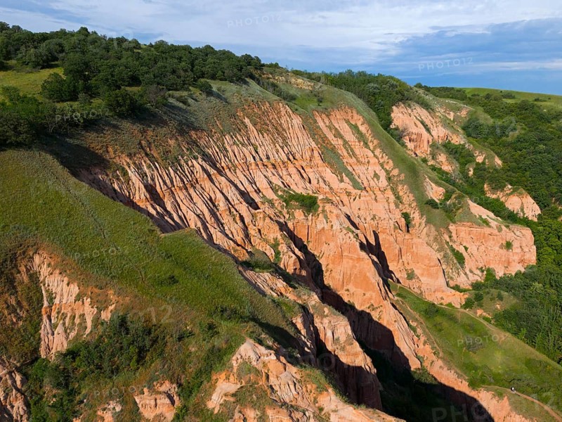 Hilly rocky landscape with traces of erosion, surrounded by green trees and a blue sky, aerial view, Râpa Ro?ie, Rapa Rosie, Red Gorge, nature reserve, Sebe?, Transylvania, Alba County, Romania