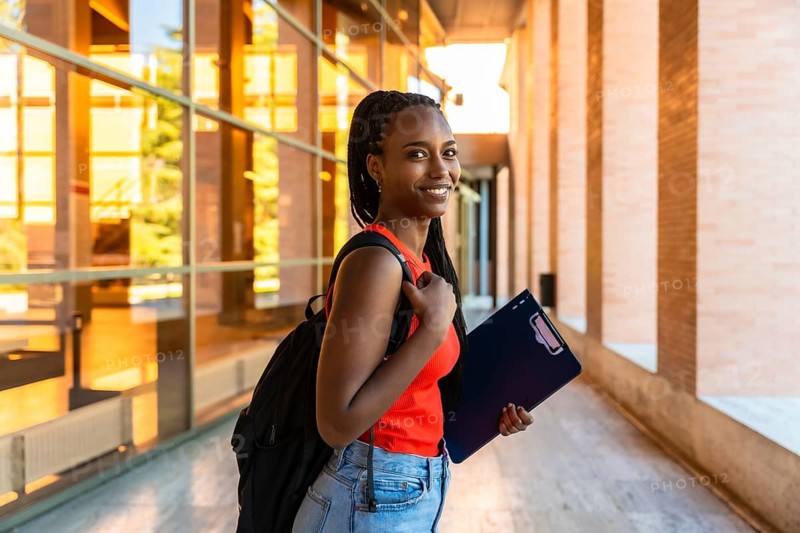 Confident young black woman student with backpack and folder smiling, walking on a modern university campus, representing education, learning, and college life