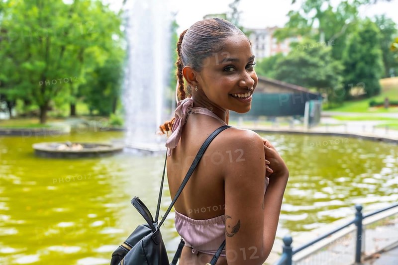 Happy young woman standing outdoors, turning her head and smiling cheerfully, enjoying a beautiful sunny day in a public park with a fountain and green foliage in the background
