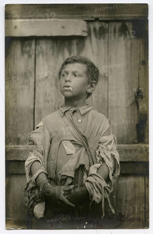 Jewish beggar, boy, Vilnius, Lithuania, poverty, hunger, hardship, orphan, cigarette, World War I, A boy in torn clothes stands in front of a wooden wall and looks seriously to the side, 1916