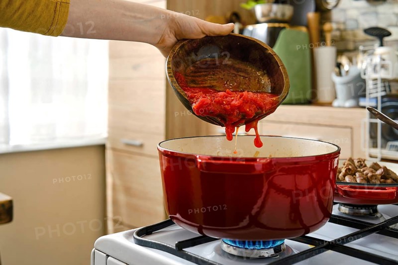 A person pours chopped tomatoes from a bowl into a red pot on a stove. The kitchen has wooden cabinets and various cooking tools. Daylight comes through the window as cooking occurs